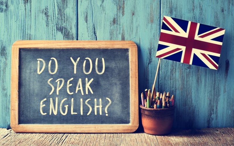 a chalkboard with the text do you speak english? written in it, a pot with pencils and the flag of the United Kingdom, on a wooden desk, with a filter effect
